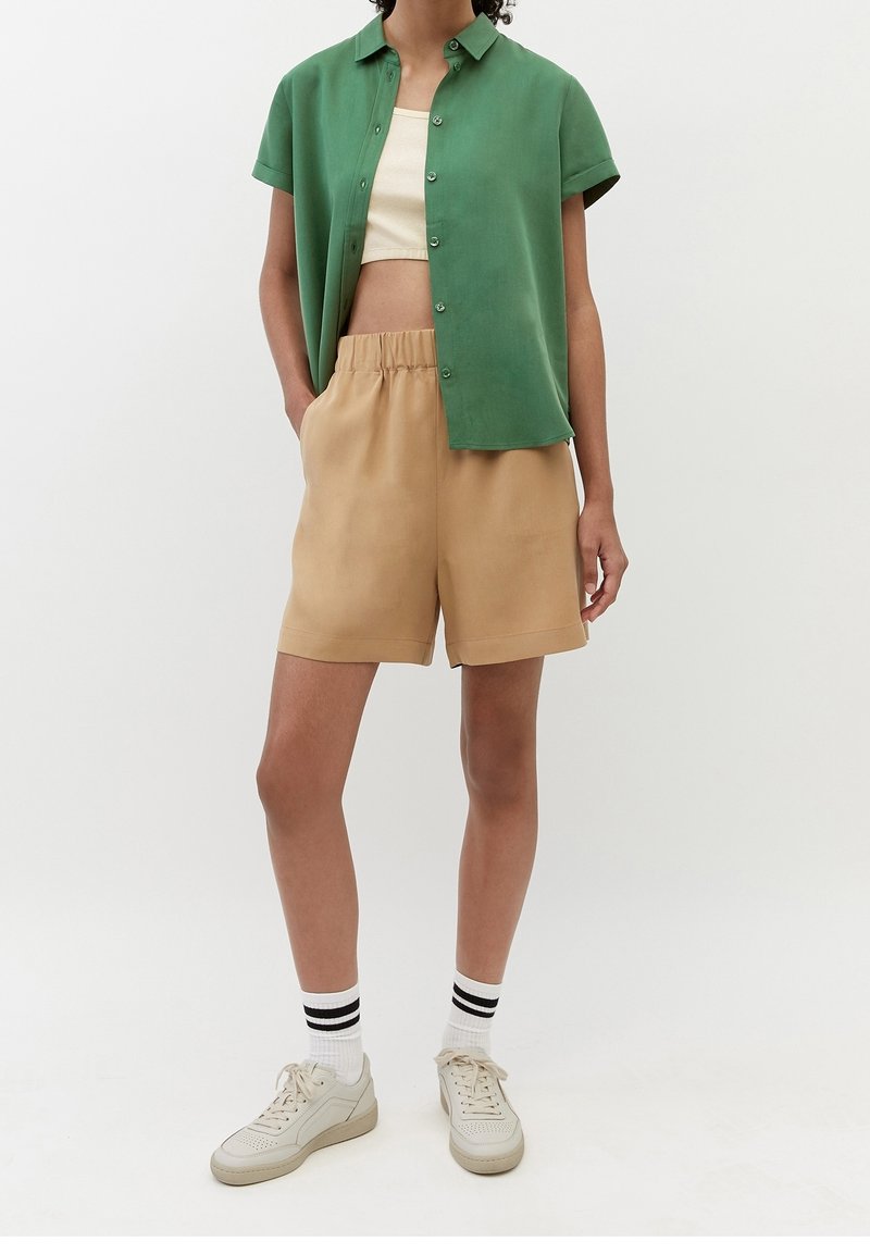 Grön kortärmad skjorta med knappar över en beige elastisk midjeshorts. Stylad med krämfärgade sneakers och vita strumpor med svarta ränder.