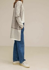 Dlouhý, světle šedý kardigan s texturovanými pruhy, nošený přes bílé tričko a modré džíny, doplněný světle barevnými teniskami.