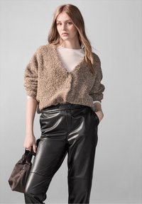Fuzzy beige cardigan met een V-hals, gedragen over een lichte top en gecombineerd met zwarte leren broek. Model houdt een donker handtas vast.