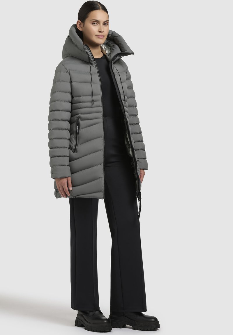 Giacca puffer grigia con colletto alto, tasche con zip e texture a coste. Abbinata a pantaloni neri a gamba larga e stivali neri chunky.