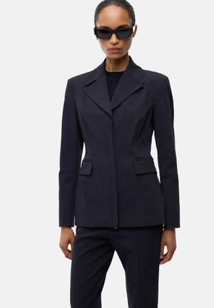 Donna con blazer nero aderente e pantaloni abbinati con occhiali da sole rettangolari neri, in piedi davanti a uno sfondo chiaro e uniforme.