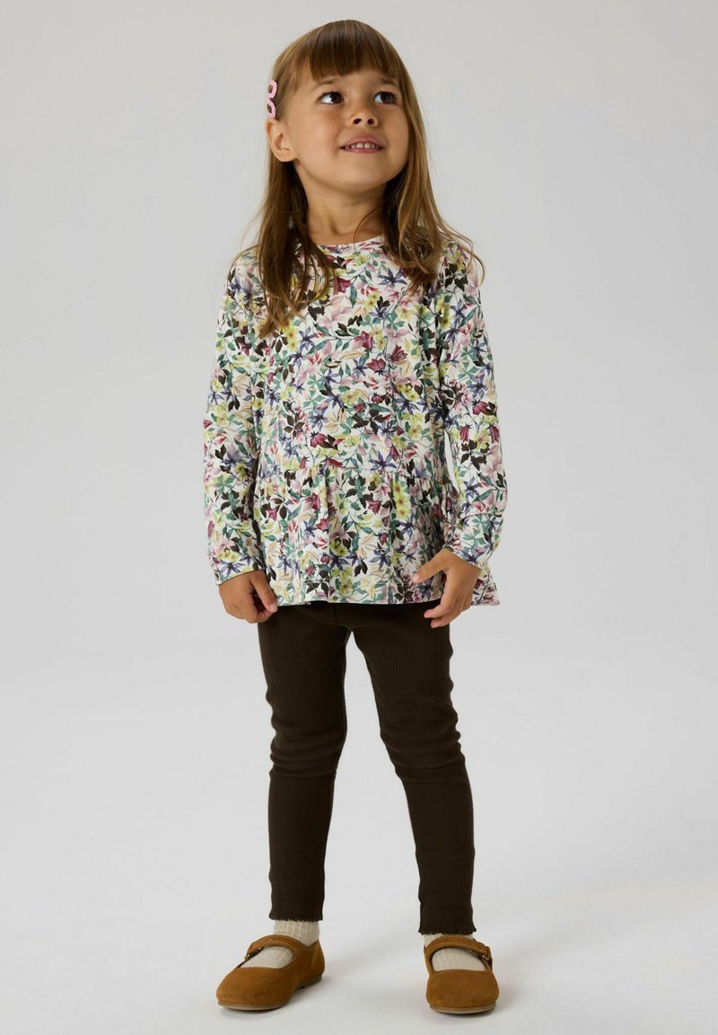 Young girl with brown hair wearing a floral long-sleeve top, dark leggings, beige socks, and brown Mary Jane shoes, looking upward.