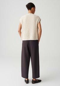 Cremfarbener, strukturierter Strickoberteil mit kurzen Ärmeln, kombiniert mit lockeren, dunklen Hosen und schwarzen Mesh-Schuhen. Schlichtes Design, neutrale Farben.