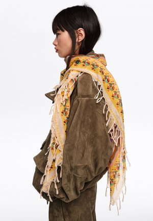 Mujer joven con chaqueta de ante verde oliva y bufanda amarilla con flecos florales sobre los hombros, mirando hacia la izquierda sobre fondo blanco.