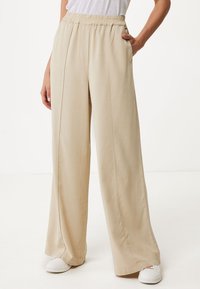Pantalons beige à jambes larges avec une taille élastique, des poches latérales et une texture lisse. Portés avec des chaussures blanches, sur un fond neutre.