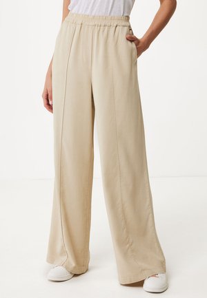 Beige wijde broek met een elastische tailleband, zijzakken en een gladde textuur. G gedragen met witte schoenen, staande op een neutrale achtergrond.