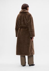 Mulher vestindo um longo casaco de pelúcia marrom por cima de calças marrons e sapatos pretos, de pé contra um fundo branco liso, de costas.