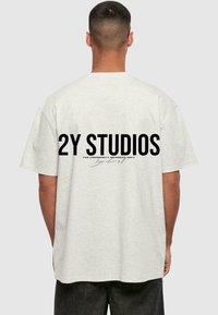 Lys grå, overdimensjonert T-skjorte med korte ermer. Har store svarte bokstaver på baksiden: "2Y STUDIOS" og mindre tekst under.