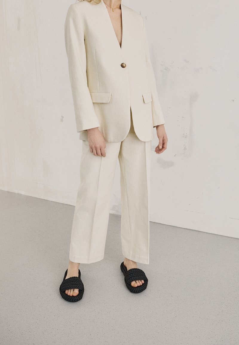 Personne portant un costume couleur crème avec un pantalon à jambes larges et un blazer boutonné, assorti à des sandales noires tissées à bouts ouverts sur un sol gris.