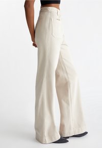 Hæl-høj, brede bukser i beige stof. Har baglommer og en glat tekstur. Kantlinjen sidder over skoene.