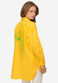 Camicia gialla a maniche lunghe con vestibilità rilassata, caratterizzata da un design grafico ricamato verde sulla parte posteriore e spacchi laterali all'orlo.