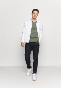 Weißes Hemdjacke über grünem Shirt, dunklen Jeans und weißen Sneakers. Einfaches Design, glatte Textur, mit Taschen an der Jacke.