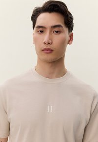 Fiatal férfi rövid sötét hajjal, bézs színű pólót visel, amelyen a mellkason középen egy fehér "II" logó látható, egyszínű világos háttér előtt.