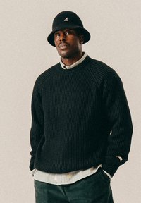 Ein Mann trägt einen schwarzen Strickpullover über einem gestreiften Shirt und einem schwarzen Bucket Hat. Er steht mit den Händen in den Taschen vor einem schlichten Hintergrund.
