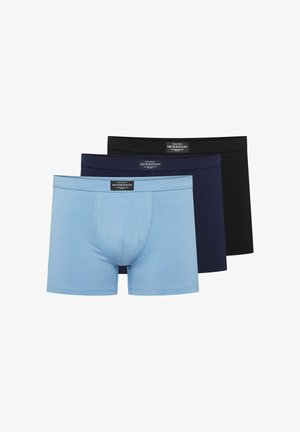 Muskelöser Mann mit kurzem Haar, der hellblaue Boxershorts trägt, steht mit einem erhobenen Arm und der Hand, die den Hinterkopf berührt.