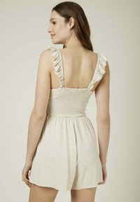 Rompers beige clair en tissu texturé, avec un corsage smocké et des bretelles à volants. Le dos est ouvert et présente une coupe courte ample.