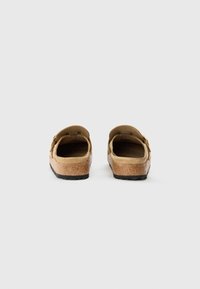 Sabots beiges avec semelle en liège et en caoutchouc noir, dotés d'une boucle dorée ajustable et d'un design à dos ouvert. Texture en suede lisse.