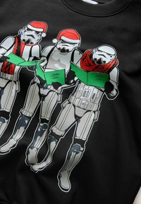 Felpa nera con tre stormtrooper in cappelli di Babbo Natale, che tengono in mano carte di Natale verdi. Il design è grafico e colorato con dettagli accattivanti.