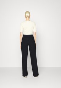 Kobieta z blond włosami w kok, ubrana w kremowy top z krótkim rękawem, czarne spodnie o wysokim stanie z szerokimi nogawkami oraz kremowe buty na kwadratowym nosku, stojąca tyłem.