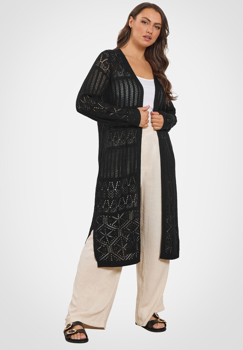 Vrouw die een lange zwarte opengebreide cardigan draagt over een wit topje, beige wijde broekspijpen en zwarte sandalen met gouden gespen, poserend staand.