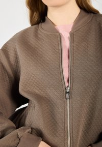 Brown quilted bomber jacket with a silver zipper, ribbed cuffs, and a pink inner lining. Features a round neck and voluminous sleeves.