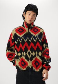 Fuzzy fleece pullover featuring a geometric pattern with red, black, and beige hues. High collar and half-zip closure; relaxed fit.