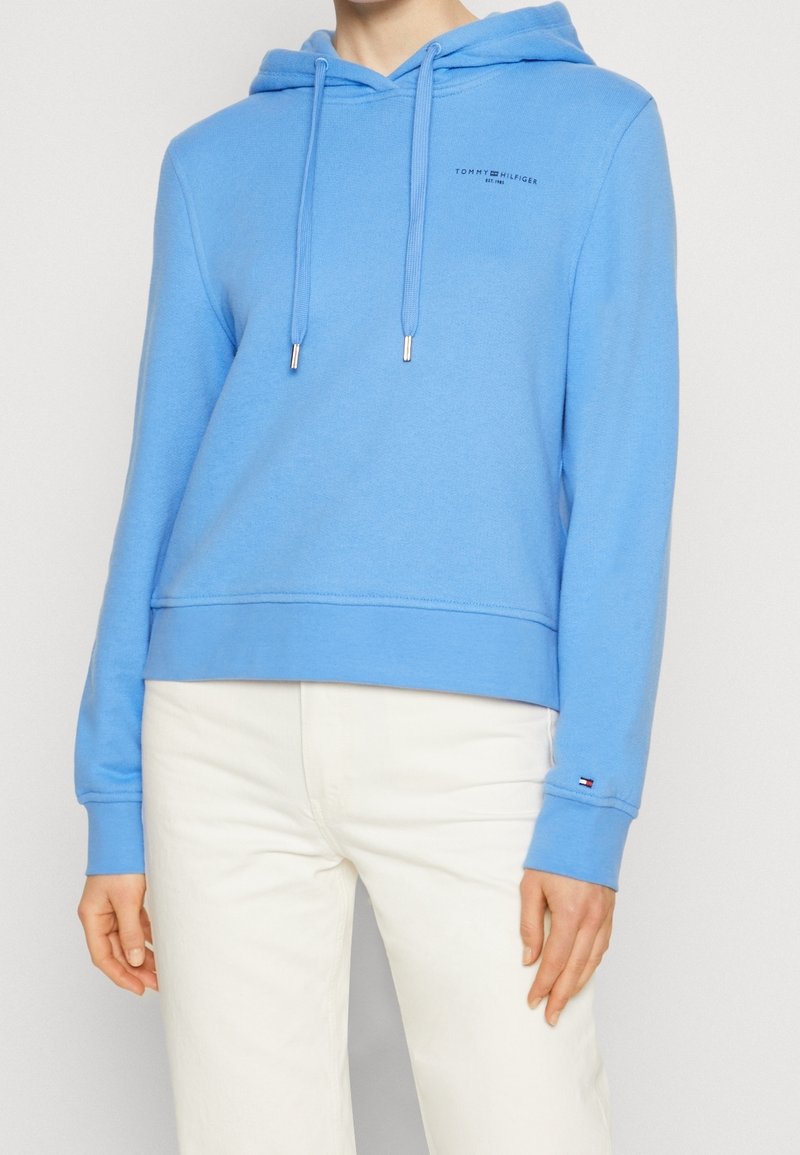 Personne portant un sweat à capuche Tommy Hilfiger bleu clair avec cordons et un pantalon blanc, montrant le haut du corps et les bras.