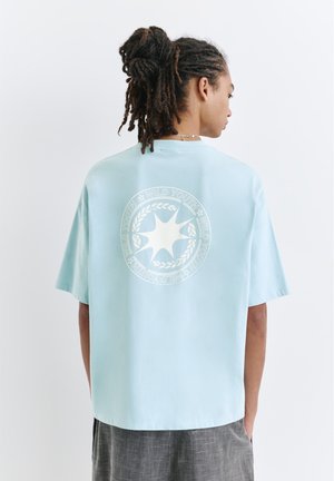 Personne avec des dreadlocks portant un t-shirt oversize bleu clair avec un logo étoile blanc "Wild Youth" dans le dos, debout devant un fond blanc.