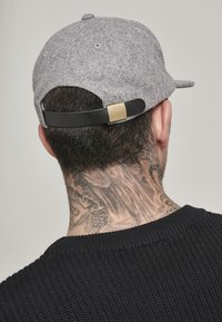 Grå følt baseballcap med en svart stropp og gullhardware bak. Viser et nærbilde av hår og en tatovering i nakken.