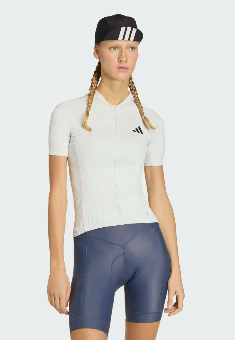 Camiseta de ciclismo gris claro con cremallera frontal y logo negro, combinada con pantalones cortos acolchados azul marino. El modelo lleva una gorra negra con rayas blancas.