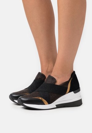 Pieds portant des baskets compensées à enfiler noires et bronze avec de larges sangles élastiques noires croisées et des semelles blanches sur un fond clair.