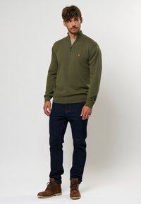 Sudadera verde oliva con cremallera y textura acanalada, combinada con jeans azul oscuro. Detalle notable: pequeño logo naranja en la sudadera. Botas marrones.