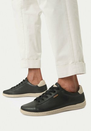 Stopy w czarnych niskich skórzanych trampkach z beżowymi podeszwami i akcentami na pięcie, w połączeniu z podwiniętymi, kremowymi spodniami.