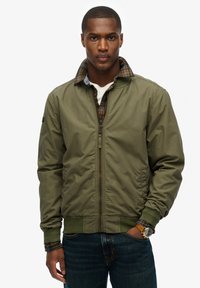 Olive green bomber jacket with a zip front, ribbed cuffs, and collar. Interior features plaid fabric. Model wearing dark jeans and a watch.