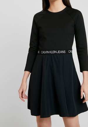 Femme portant une robe noire à manches longues avec une ceinture élastique marquée « Calvin Klein Jeans », debout devant un fond gris uni.