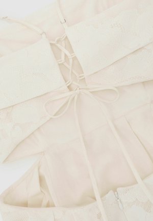 Koronkowy biustonosz bralet w kremowym kolorze z cienkimi ramiączkami i detalem zawiązywania na plecach, ułożony na płasko na tle jasnej, pasującej tkaniny.