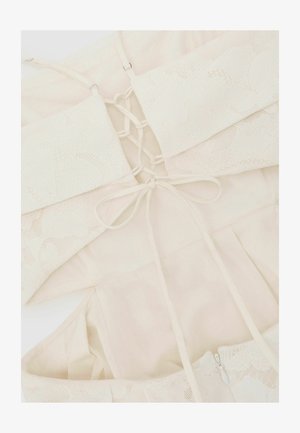 Koronkowy biustonosz bralet w kremowym kolorze z cienkimi ramiączkami i detalem zawiązywania na plecach, ułożony na płasko na tle jasnej, pasującej tkaniny.