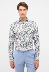 Witte shirt met kleurrijke paisley- en bloemmotieven in blauw, geel en groen; lange mouwen; gecombineerd met lichtblauwe broeken.
