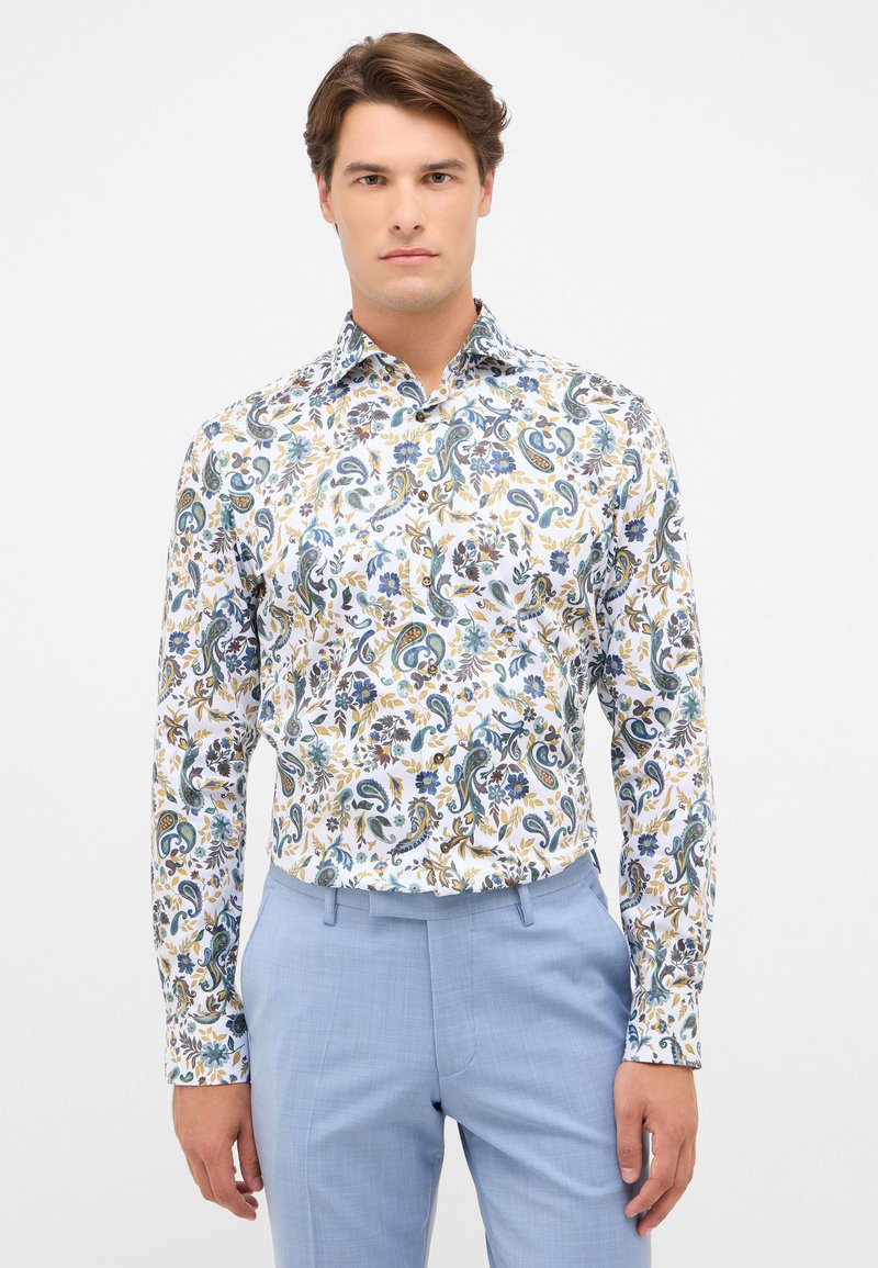 Witte shirt met kleurrijke paisley- en bloemmotieven in blauw, geel en groen; lange mouwen; gecombineerd met lichtblauwe broeken.