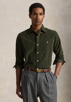 Jeune homme debout, une main dans la poche, portant une chemise boutonnée vert foncé et un pantalon plissé gris avec une ceinture marron.