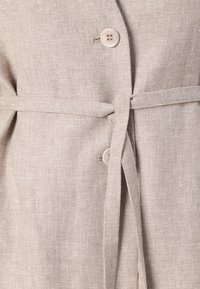 Beige liinablazer, jossa on solmittava vyö, sisältää kaksi nappia ja teksturoitua kangasta. Korostaa toiminnallista muotoilua ja minimalistista tyyliä.