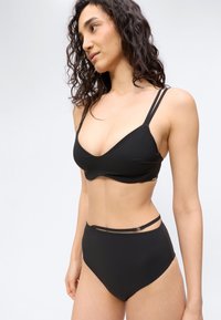 Γυναίκα με μακριά σγουρά μαλλιά που φοράει μαύρο μπικίνι δύο τεμαχίων με διπλούς ιμάντες στο πάνω μέρος και σλιπ ψηλόμεσο με λεπτομέρεια κοψίματος.