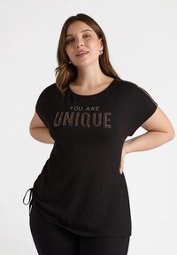Camiseta negra de manga corta con la frase "ERES ÚNICO" en strass, con escote en forma de lágrima y lazos laterales ajustables.