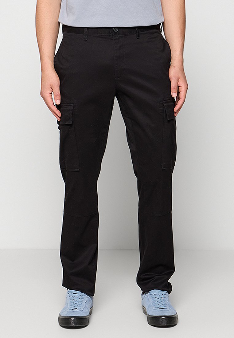 Homme portant un pantalon cargo noir avec des poches à rabat sur les cuisses et des baskets bleu clair, se tenant devant un fond clair.