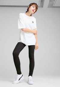Persona joven vistiendo una camiseta blanca de Puma, leggings negros y zapatillas de plataforma blancas, de pie contra un fondo gris liso.