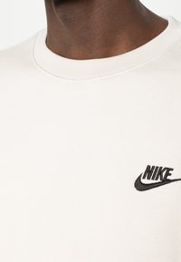 Sudadera de felpa crema con un cuello redondo acanalado. Presenta un pequeño logo de Nike bordado en negro en el pecho. Textura suave.