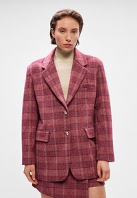 Red plaid blazer with a notched collar, front pockets, and two button closure. Fabric has a textured feel with contrasting lines.