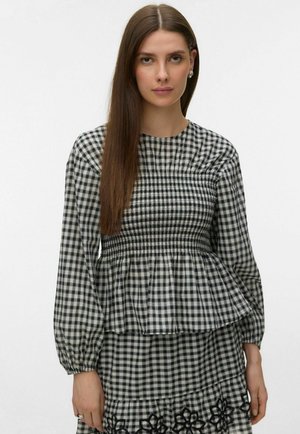 Femme aux cheveux longs et raides portant un chemisier à carreaux noirs et blancs et une jupe assortie avec une broderie florale noire.