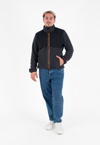 Zwarte en grijze jack met rits en hoge kraag, textuurstof en contrasterende oranje rits. Gekoabd over blauwe denim jeans en witte sneakers.