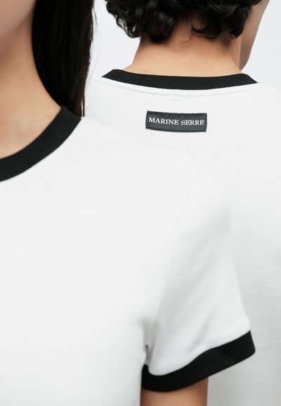 Λευκό μπλουζάκι με μαύρο περίγραμμα και μαλακή υφή. Μια ετικέτα στο πίσω μέρος εμφανίζει το "MARINE SERRE" σε λευκό σε μαύρο φόντο.
